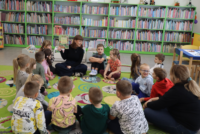 Na zdjęciu znajduje się 16 przedszkolaków i 2 kobiety na dywanie, którzy siedzą w okręgu, kobieta na przeciwko pokazuje dzieciom książkę o warzywach. Po prawej stronie znajdują się małe stoliki, a w tle półki z książkami.