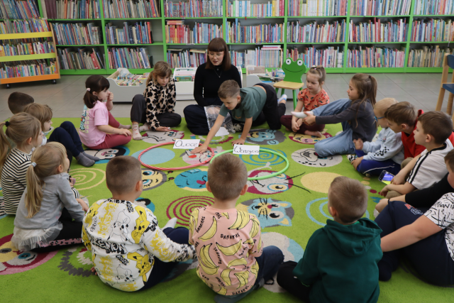 Na zdjęciu znajdują się dzieci na dywanie, siedzą w okręgu, na przeciwko nich siedzi kobieta przed nią znajdują się dwa hula hop, w jeden znajduje się napis owoce, w drugiej warzywa. Chłopiec siedzący po prawo obok kobieta układa ilustrację w odpowiednią obręcz. W tle znajdują się półki z książkami.