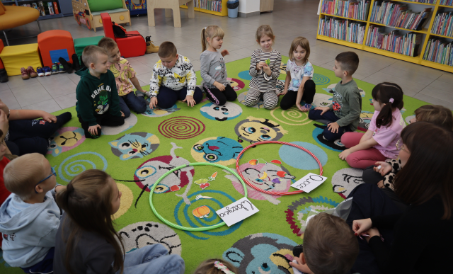 Na zdjęciu znajdują się przedszkolaki wraz z kobietą na dywanie, po środku leżą dwa hula hop z kartkami owoce i warzywa oraz odpowiednio posegregowanymi ilustracjami. W tle znajdują się półki z książkami oraz pufy ułożone w pociąg.