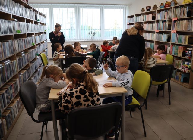 Na zdjęciu znajdują się dzieci przy stolikach i kolorują koszyczki na kartkach, między stolikami stoją dwie kobiety. W tle znajdują się półki z książkami oraz okna.