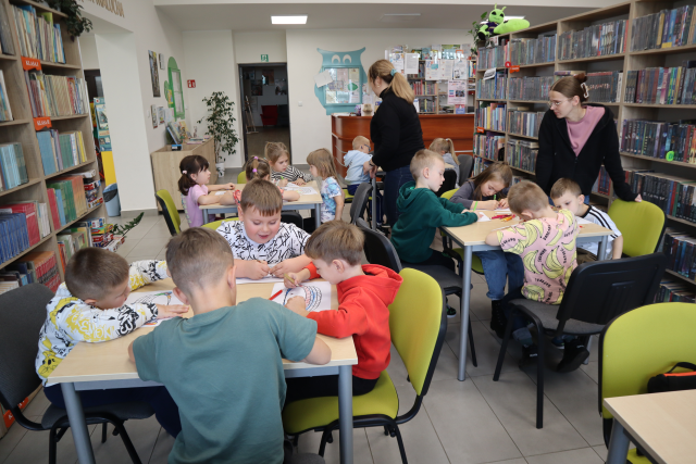 Na zdjęciu znajduje się 16 przedszkolaków przy 4 stolikach, przy których kolorują koszyczki na kartce. Między stolikami stoją dwie kobiety. W tle znajdują się półki z książkami, drzwi, biurko biblioteczne.