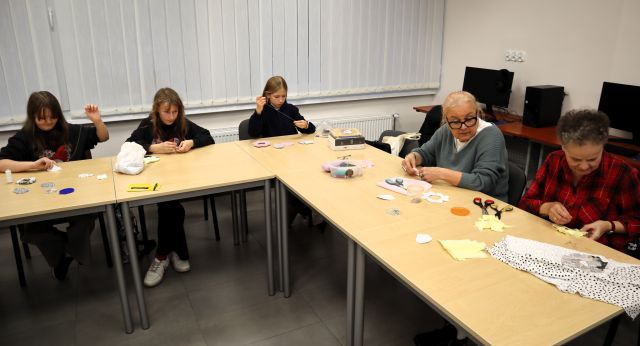 Na zdjęciu znajdują się 3 dziewczynki i dwie dorosłe kobiety przy złączonych stolikach w literę L, na stolikach znajdują się materiały, nici, z których uczestniczki szyją ośmiorniczki. Warsztaty odbywają się w sali komputerowej.