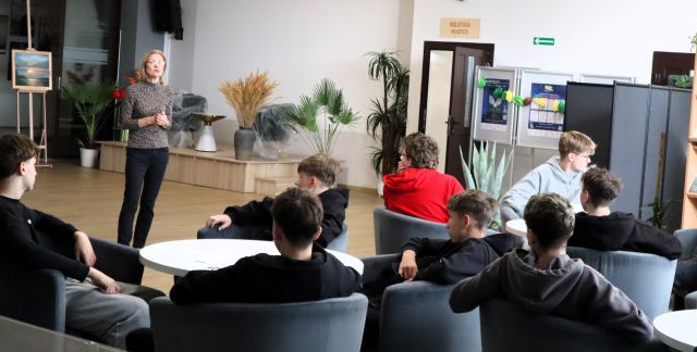 Na zdjęciu znajduje się młodzież przy okrągłych stolikach, na fotelach, a przed nimi stoi kobieta na holu biblioteki. W tle znajdują się czarne tablice z wystawką, drzwi, rośliny i mała scena.