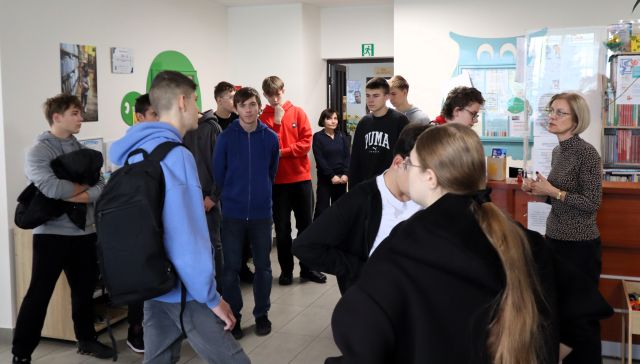 Na zdjęciu znajduje się młodzież w sali bibliotecznej, po prawej stronie przy biurku stoki kobieta. W tle znajdują się drzwi, a po lewej stronie fotografie na ścianach.