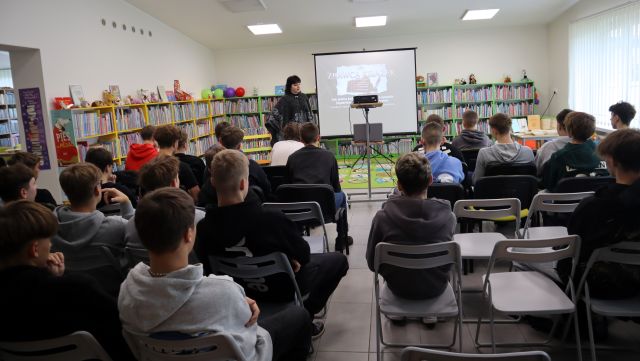 Na zdjęciu znajduje się około 40 młodzieży na krzesłach, przed nimi stoi kobieta przy ekranie, na którym wyświetlana przez projektor. W tle znajdują się półki z książkami.