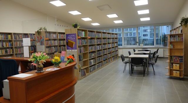 z lewej strony część brązowej lady bibliotecznej, w tle regały z książkami, po prawej stronie stoliki z krzesłami