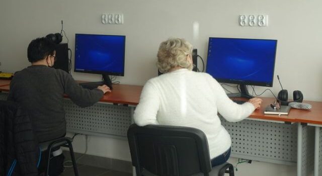 dwie kobiety odwrócone tyłem siedzą przy komputerach, które stoją na brązowych stołach. Jedna kobietaw białej bluzce, a druga w czarnej