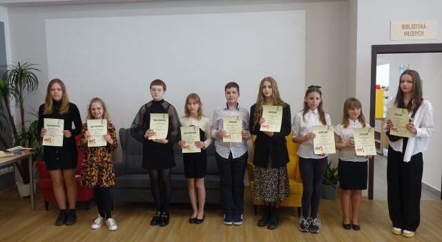 na tle białej ściany stoi 8 dziewczynek i pośrodku jeden chłopiec w białej bluzce w kropki. Dziewczynki z lewej strony mają białe bluzki, a z prawej ciemne. W dłoniach trzymają dyplomy