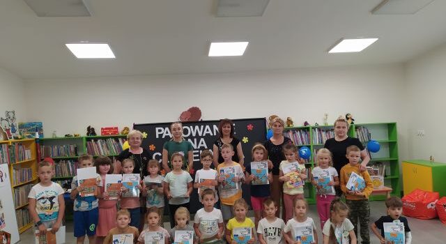 w pierwszym rzędzie dzieci siedzą na zielonym dywanie, na nimi stoją dzieci, za nimi 4 kobiety, za n imi czarna tablica