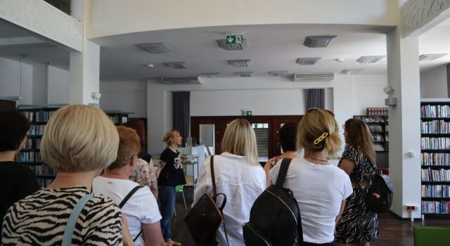 kobiety stoją tyłem, przed nimi kobieta opowiadająca na tle białej ściany