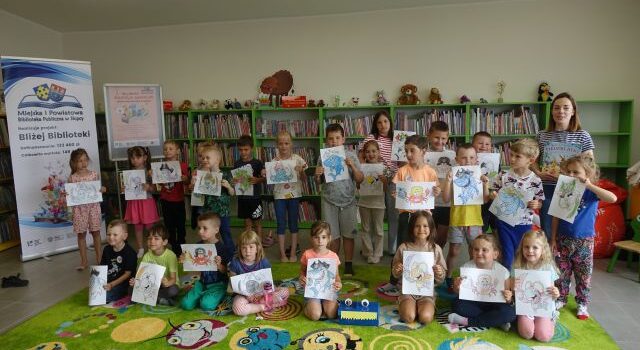 w pierwszym rzędzie dzieci siedzą na zielonym dywanie, w drugim i trzecim rzędzie stoją. Wszyscy trzymają w rękach kartki papieru z malowankami. Za nimi zielone regały z książkami