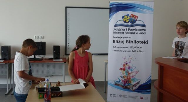 chłopiec siedzi przy brązowym stole i rozkłada planszę z szachami, dziewczynka stoi, w drzwiach stoi chłopiec, za nimi duży plakat z nazwą zadania i logo biblioteki