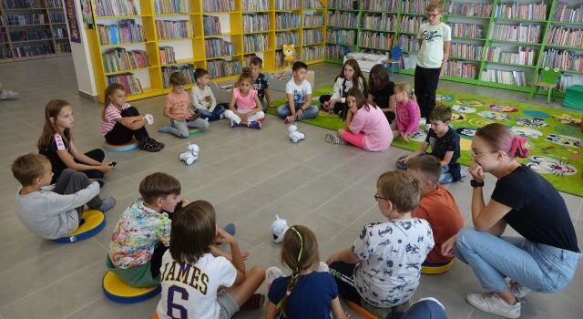 dzieci siedzą w kole na siwej podłodze, przed nimi białe małe roboty, za nimi zielone i żółte regały z książkami