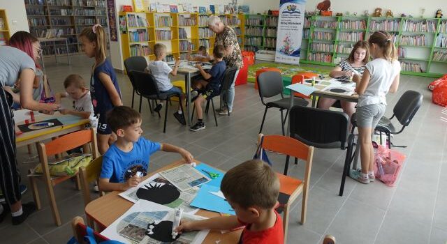 przy stołach siedzą dzieci i naklejają wycięte z kartek zęby rekina, za nimi żółte i zielone regały z książkami
