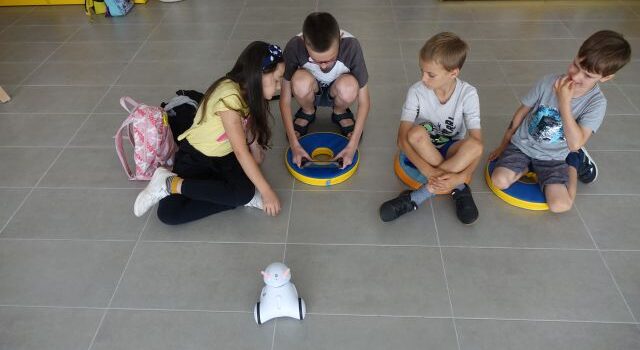 na podłodze siedzą dzieci, chłopiec w dłoniach trzyma tablet, przed nimi jeździ biały mały robot