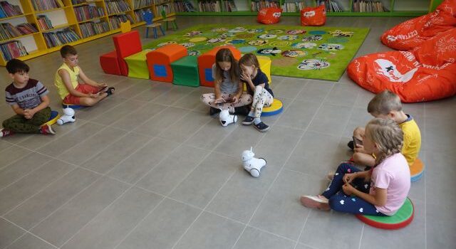 na podłodze siedzą dzieci i sterują małymi białymi robotami, za nimi zielony dywan, kolorowe pufy, za nimi zielone i żółte regały z książkami