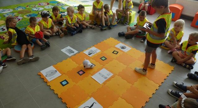dzieci w zielonych kamizelkach siedzą wokół rozłożonej na podłodze pomarańczowej macie, po której jeździ robot, którym steruje z tabletu chłopiec