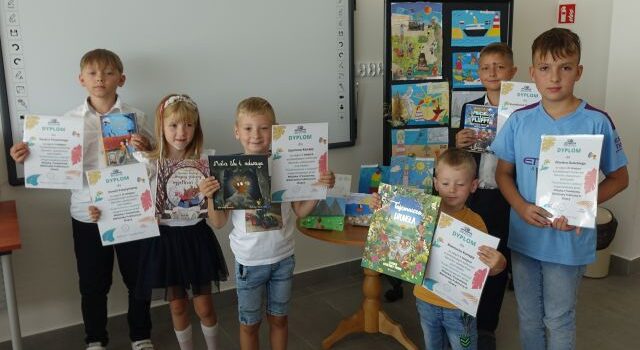 małę dzieci w jasnych bluzkach trzymają w rękach dyplomy i książki, za nimi na tablicy prace plastyczne kolorowe