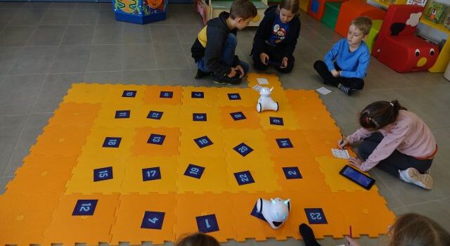 dzieci klęczą wokół pomarańczowych puzli, na których stoją małe białe roboty photon i kartki z liczbami