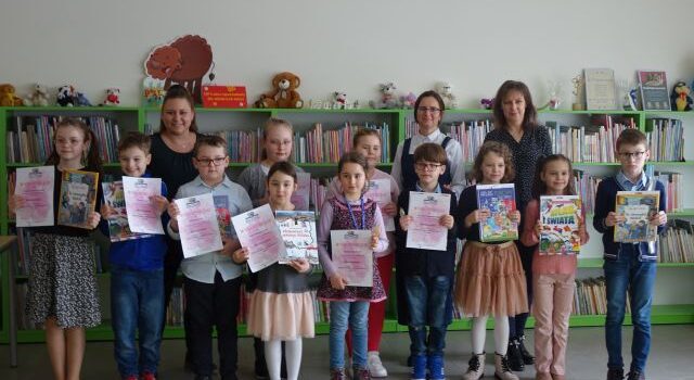 11 dzieci nagrodzonych w konkursie stoi z nagrodami książkowymi i dyplomami i pozuje do zdjęcia z nimi stoją 3 kobiety na tle regałów z książkami