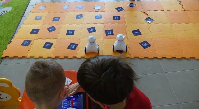 dzieci siedzą wokół pomarańczowej maty z puzli, na niej kartki z liczbami i roboty photon