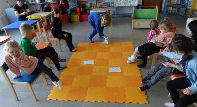 8 dzieci siedzi na krzesełkach, przed nimi na podłodze rołożona pomarańczowa mata, a na niej kartki i 2 roboty Photon