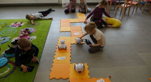 dzieci siedzą na podłodze i sterują przy pomocy tabletów robotami jeżdżącymi po pomarańczowej macie