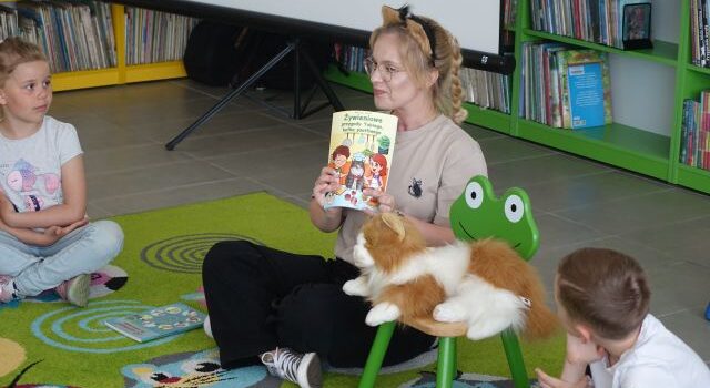 kobieta siedzi na dywanie i pokazuje dzieciom książkę, obok na krześle siedzi maskotka kota