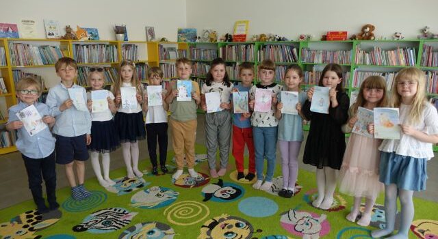 dzieci stoją na kolorowym dywanie i prezentują wykonane przez siebie prace, w tle regały z książkami