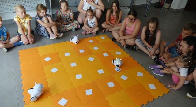 dzieci sterują przy użyciu tabletów robotami jeżdżącymi po pomarańczowej macie