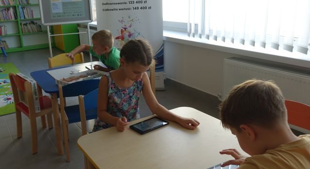 dzieci siedzą przy stolikach i sterują za pomocą tabletów robotami Photon