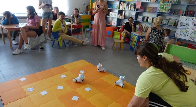 dziewczyna patrzy na roboty na pomarańczowej macie, przed nią przy stolikach siedzi młodzież