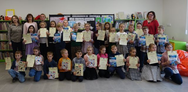 mali laureaci konkursu pozują do zdjęcia z jurorkami i bibliotekarkami oraz dyplomami i książkami, w tle wystawka z pracami dzieci