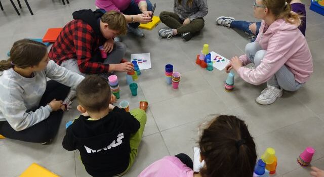 dzieci i młodzież siedzą na podłodze i ukłądają kolorowe kubeczki