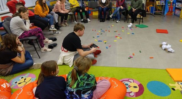 młodzież i dzieci siedzą na krzesłach i podłodze i sterują przy użyciu tabletów robotami Photon