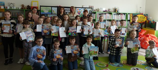 dzieci pozują do zdjęcia z dyplomami i książkami, w tle prace plastyczne wykonane przez dzieci