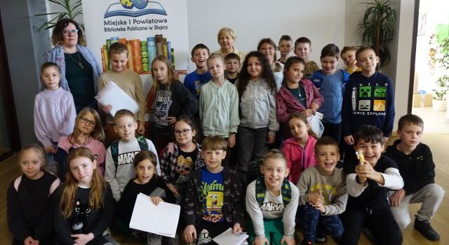dzieci z dwoma paniami pozują do zdjęcia na tle baneru z nazwą biblioteki