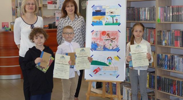 dwóch chłopców, dziewczynka i dwie bibliotekarki pozują do zdjęcia. Dzieci trzymają w rękach dyplomy i książkę, w tle rysunki dzieci