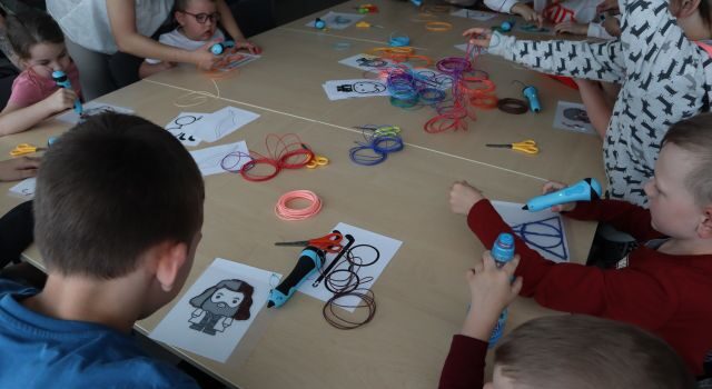 przy stołąch siedzą dzieci, na stołach długopisy 3d, filamenty i kartki z postaciami z książki o Harrym Potterze