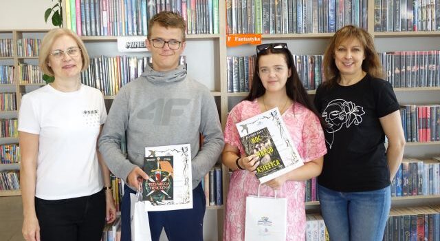 na zdjęciu z prawej strony dyrektorka biblioteki, z lewej bibliotekarka, w środku chłopak i dziewczyna z nagrodami, w tle regały z książkami