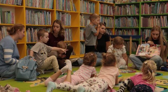 dzieci siedzą i leżą na dywanie, przed nimi siedzi bibliotekarka z książką, w tle regały z książkami