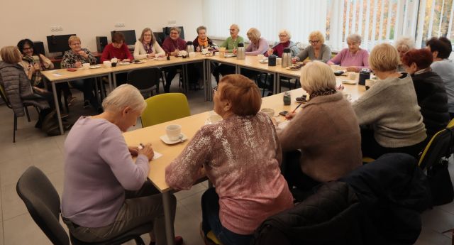 20 seniorek siedzi przy stołach ustawionych w literę u i pisze