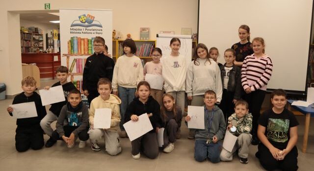 18 dzieci uczestniczących w warsztatach pozuje do zdjęcia z kartkami i ilustratorką