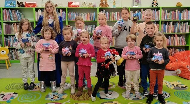 dzieci i bibliotekarka w stroju księżniczki pozują do zdjęcia ze swoimi słoikami z miasteczkami smerfów, w tle regały z książkami