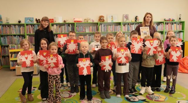 przedszkolaki pozują do zdjęcia z wykonanymi przez siebie orzełkami na czerwonej kartce, w tle regały z książkami