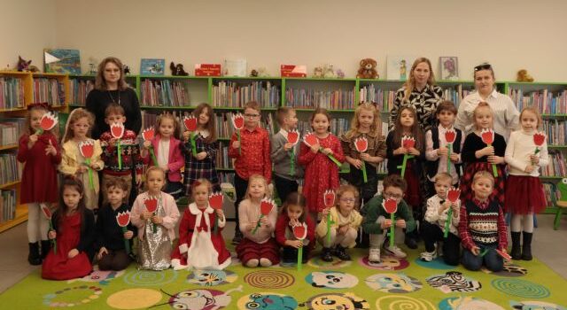 przedszkolaki, bibliotekarka i nauczycielki pozują do zdjęcia, dzieci trzymają w rękach zrobione przez siebie tulipany z kartek, w tle regały z książkami