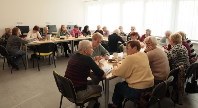 około 20 seniorek siedzi przy ustawionych w literę u stołach, rozwiązują zadania na kartkach