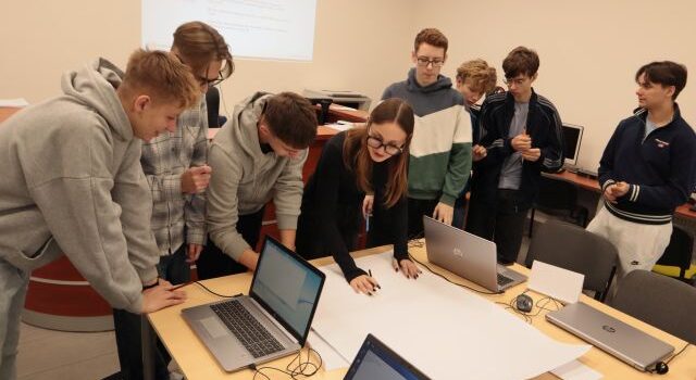 młodzież pochylona nad dużym arkuszem papieru, dziewczyna rysuje na nim, na stołach laptopy