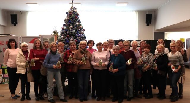 Na zdjęciu znajdują się dwadzieścia dwie kobiety stojące na tle choinki i trzymające w dłoniach pierniczki w kształcie choinki. W tle znajdują się okna.