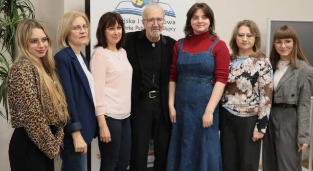 Na zdjęciu znajduje się sześć kobiet, a między nimi po środku stoi Krzysztof Daukszewicz, za nimi stoi baner z logiem biblioteki.